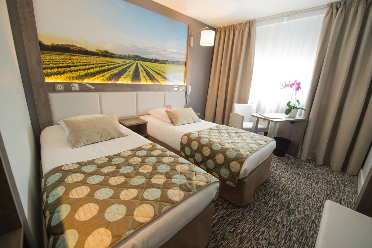A hotel room with two beds, a desk, a chair, a window with curtains, and a wall-mounted TV displaying a field.