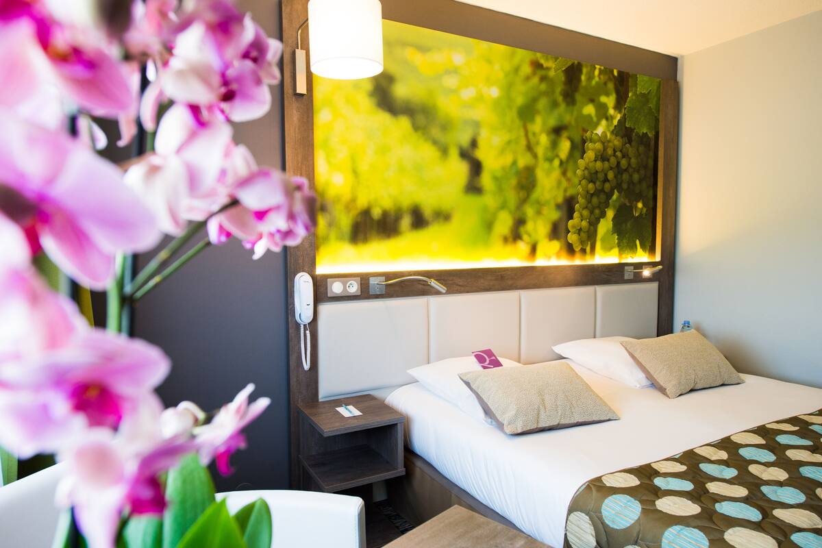 A hotel room with a bed, a nightstand, a phone, and a wall-mounted TV displaying a vineyard scene.