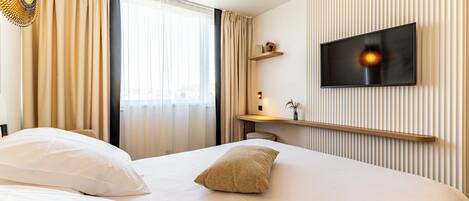 A hotel room with a large bed, a TV mounted on the wall, a bedside table with a lamp, and a window with curtains.