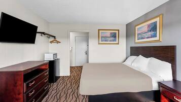 A hotel room with a large bed, a wooden dresser, a flat-screen TV mounted on the wall, a painting above the bed, and a door leading to another room.