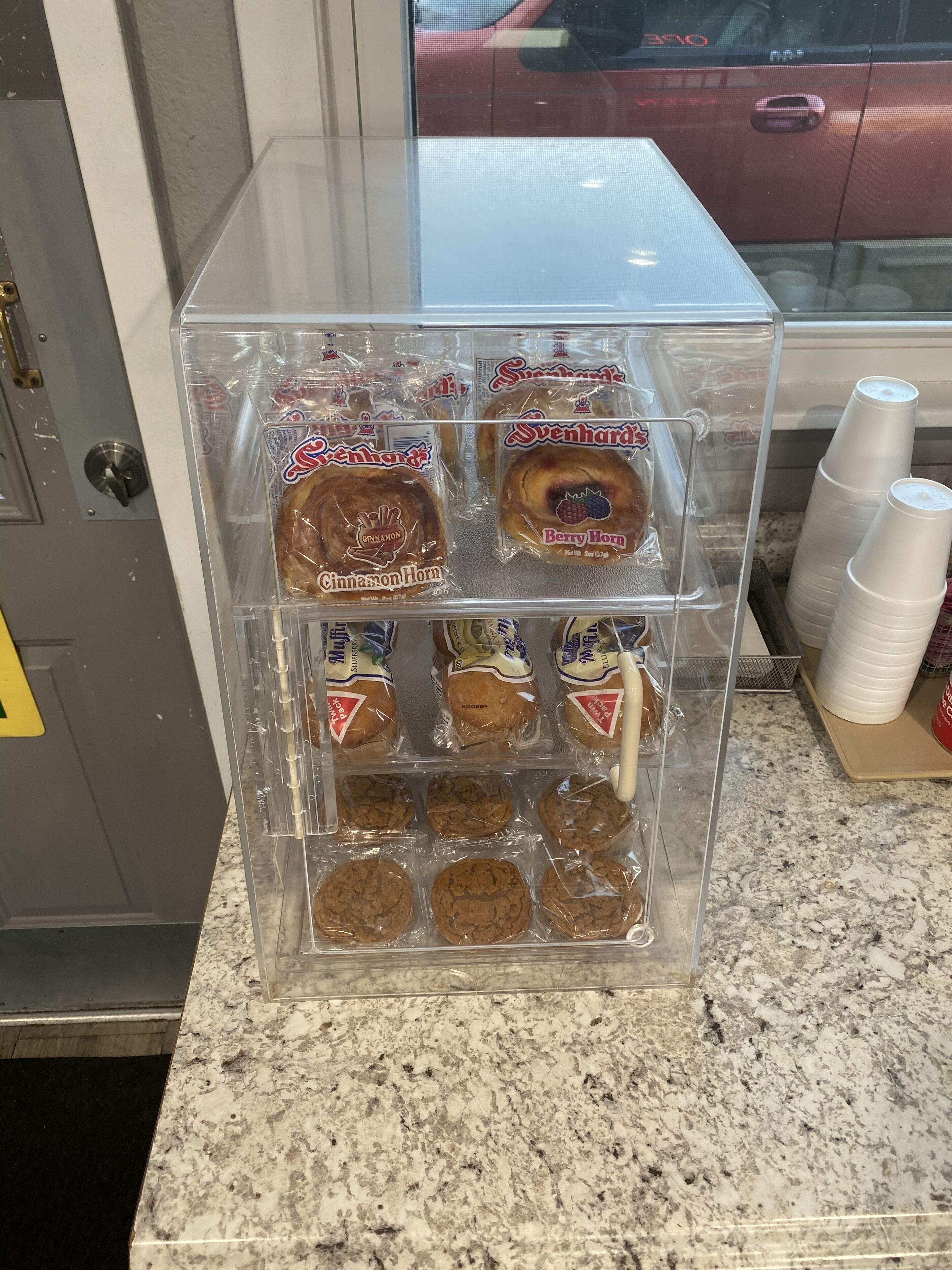 A clear plastic display case containing various packaged baked goods, including cinnamon and blueberry muffins, and cookies.