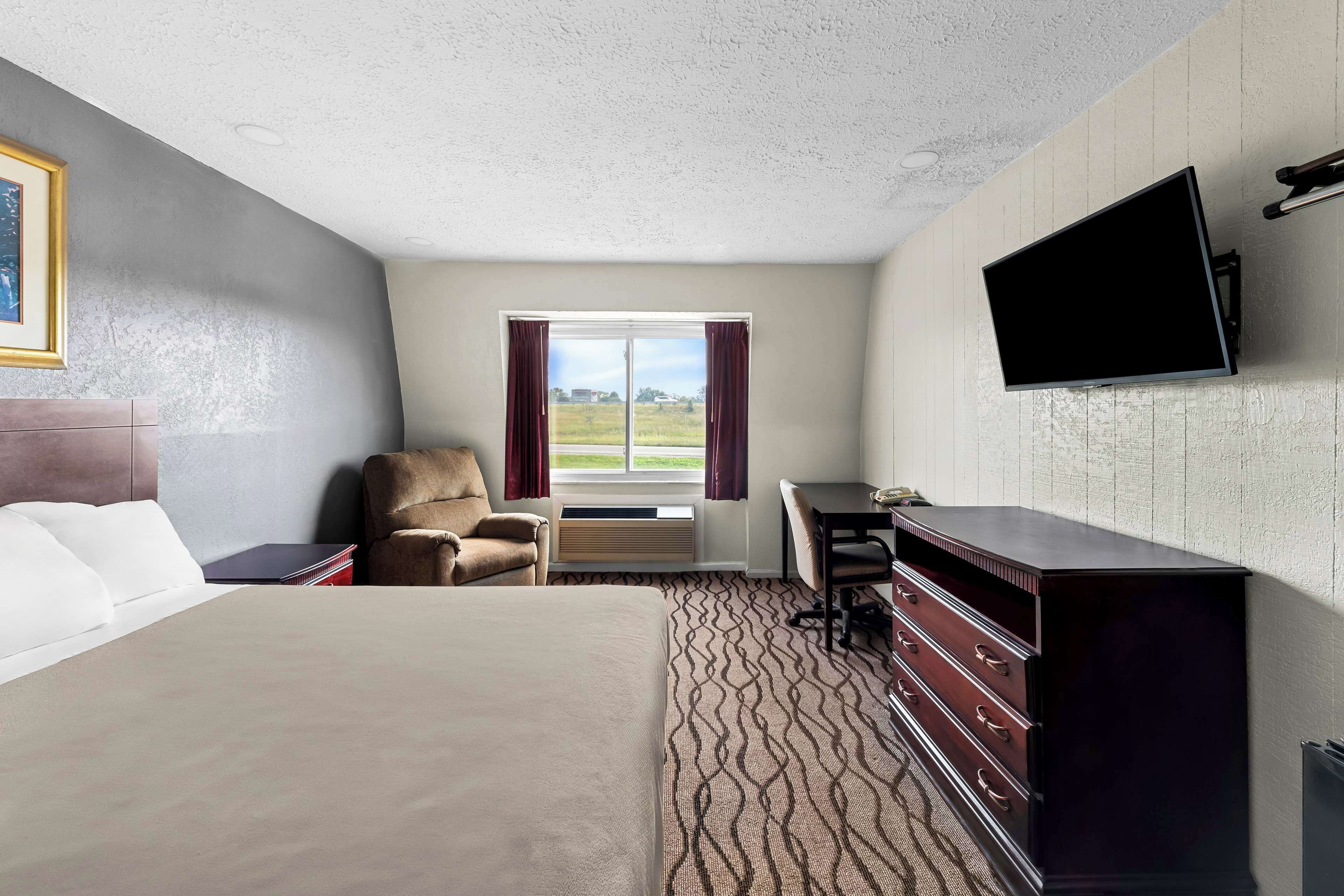 A hotel room with a bed, a desk, a chair, a TV mounted on the wall, and a window with curtains.