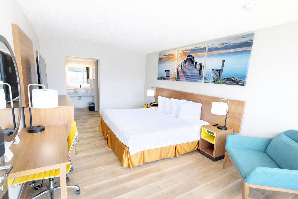 A hotel room with a bed, desk, chair, and a painting of a pier.