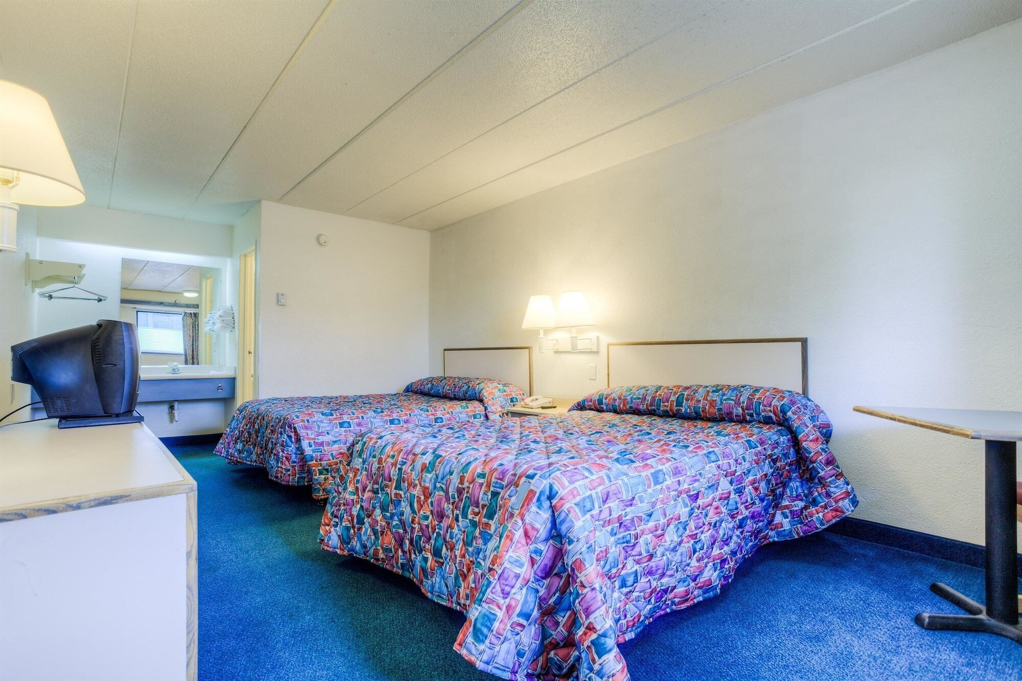 A hotel room with two beds, a desk, a chair, a lamp, and a window.