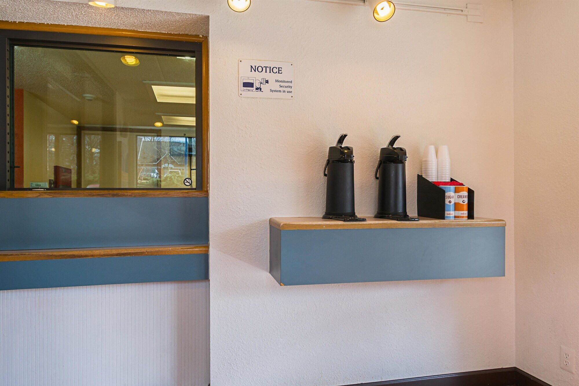 Una zona de recepción con un cartel de 'NOTICE' que indica un sistema de seguridad monitoreado. Hay dos botellas de agua negra y una cafetera en un estante.