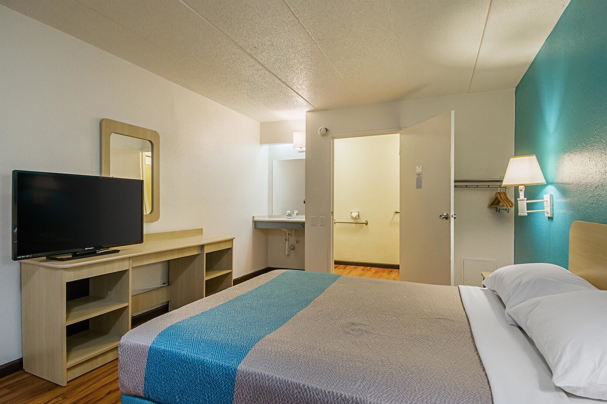 A hotel room with a bed, a TV on a wooden stand, a bathroom with a mirror, and a teal accent wall.