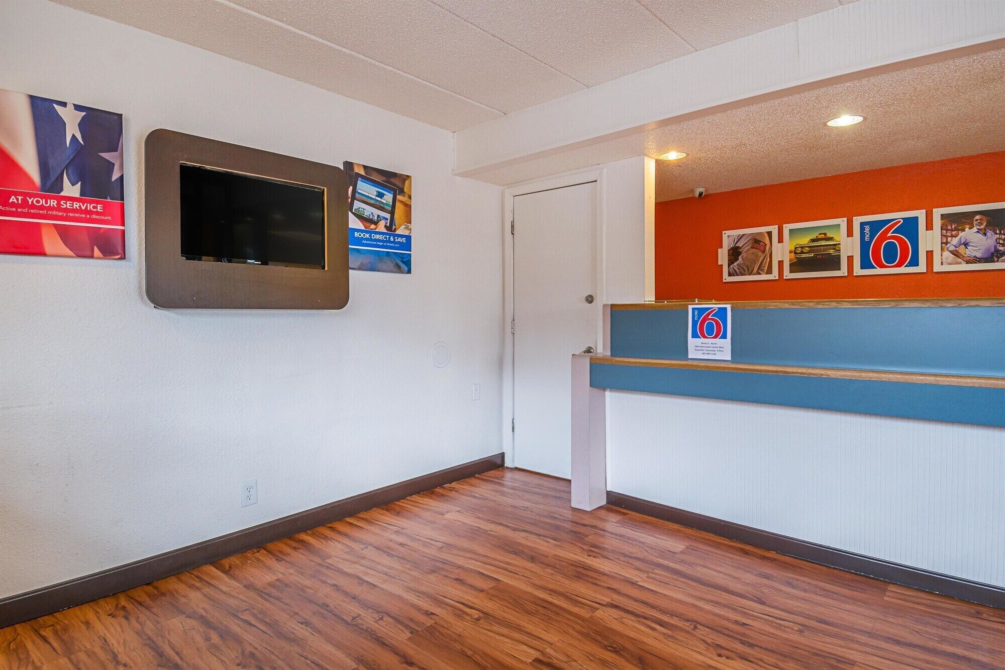 Un área de recepción con suelo de madera, un mostrador de recepción, un televisor montado en la pared y varios carteles, incluyendo uno de un motel.