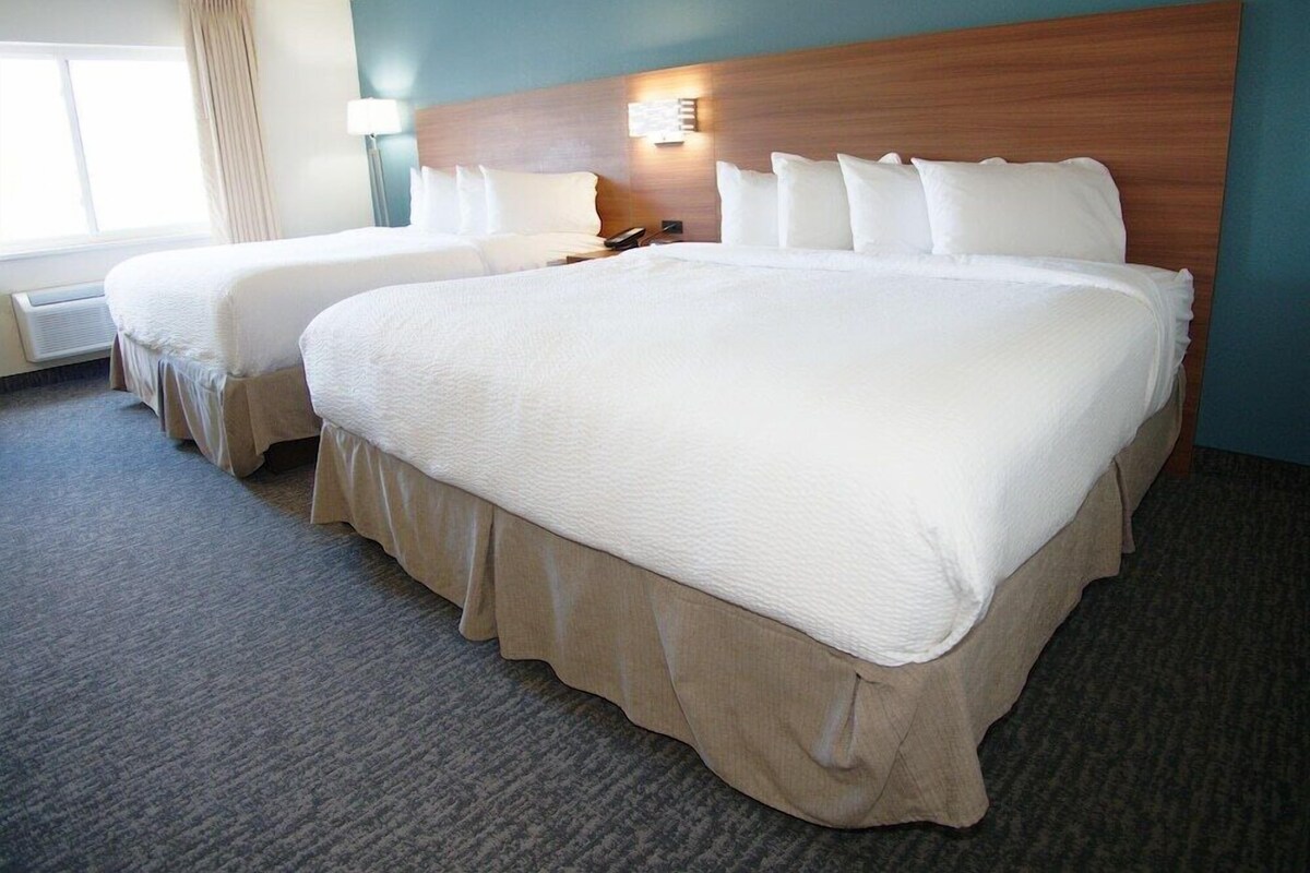 Une chambre d’hôtel avec deux lits, une tête de lit, une table de chevet, une lampe et une fenêtre avec des rideaux.