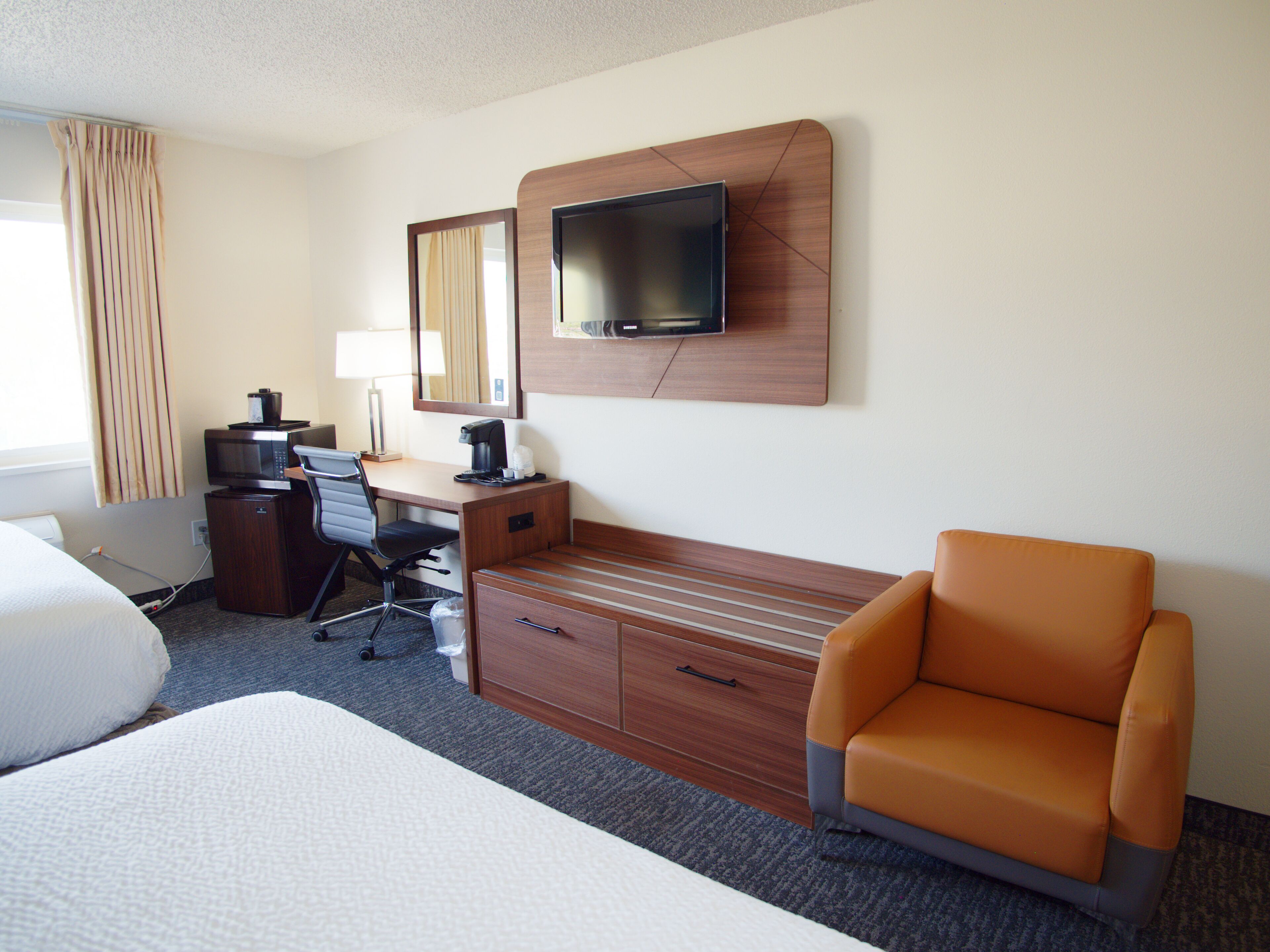 A hotel room with a bed, a desk with a computer, a coffee maker, a TV mounted on a wooden panel, and an orange armchair.