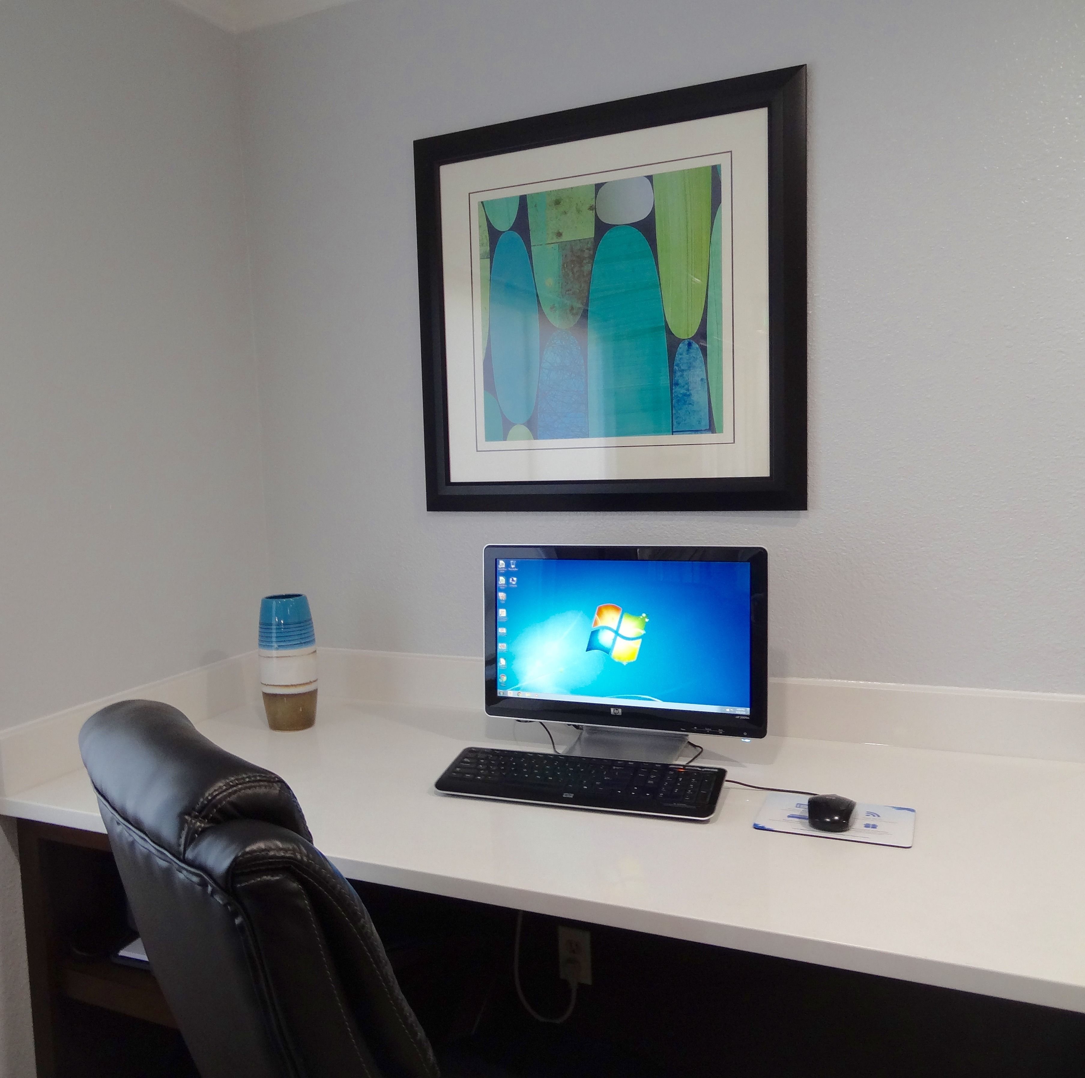 Un espace de travail avec un écran d’ordinateur, un clavier et une souris sur un bureau blanc. Un tableau abstrait encadré est accroché au mur, au-dessus du bureau.