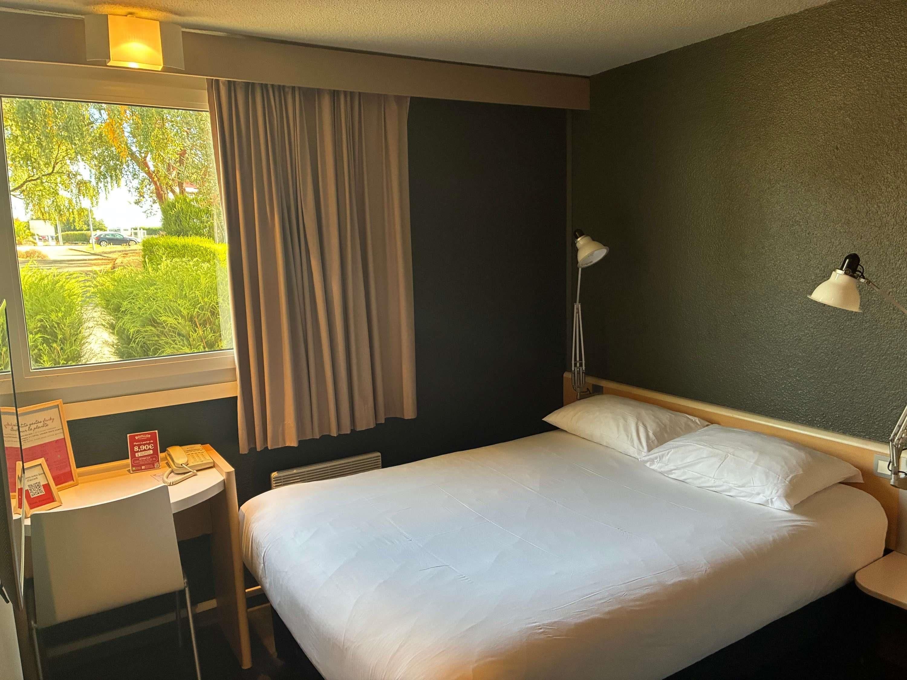 A hotel room with a bed, a desk, a chair, a window with curtains, and a lamp.