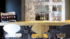 Zona de bar con estantes llenos de botellas y vasos, una barra de madera y una pared con ilustraciones en blanco y negro.