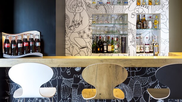 A bar area with shelves of bottles and glasses, a wooden counter, and a wall with black and white illustrations. - Ibis Lorient-Caudan (Caudan)