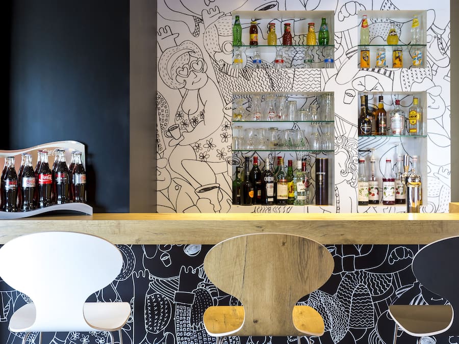 Zona de bar con estantes llenos de botellas y vasos, una barra de madera y una pared con ilustraciones en blanco y negro.