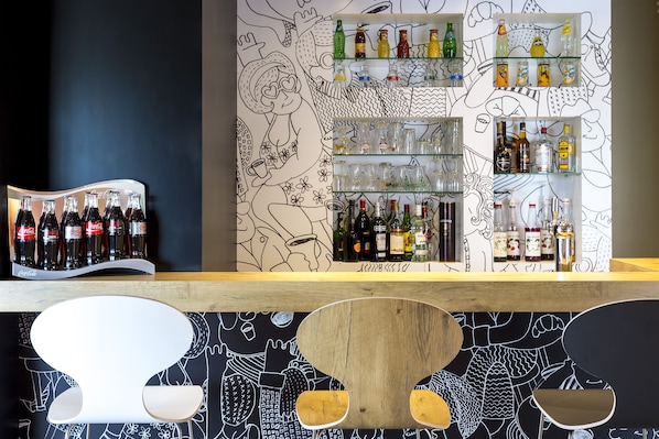 A bar area with shelves of bottles and glasses, a wooden counter, and a wall with black and white illustrations. - Ibis Lorient-Caudan (Caudan)