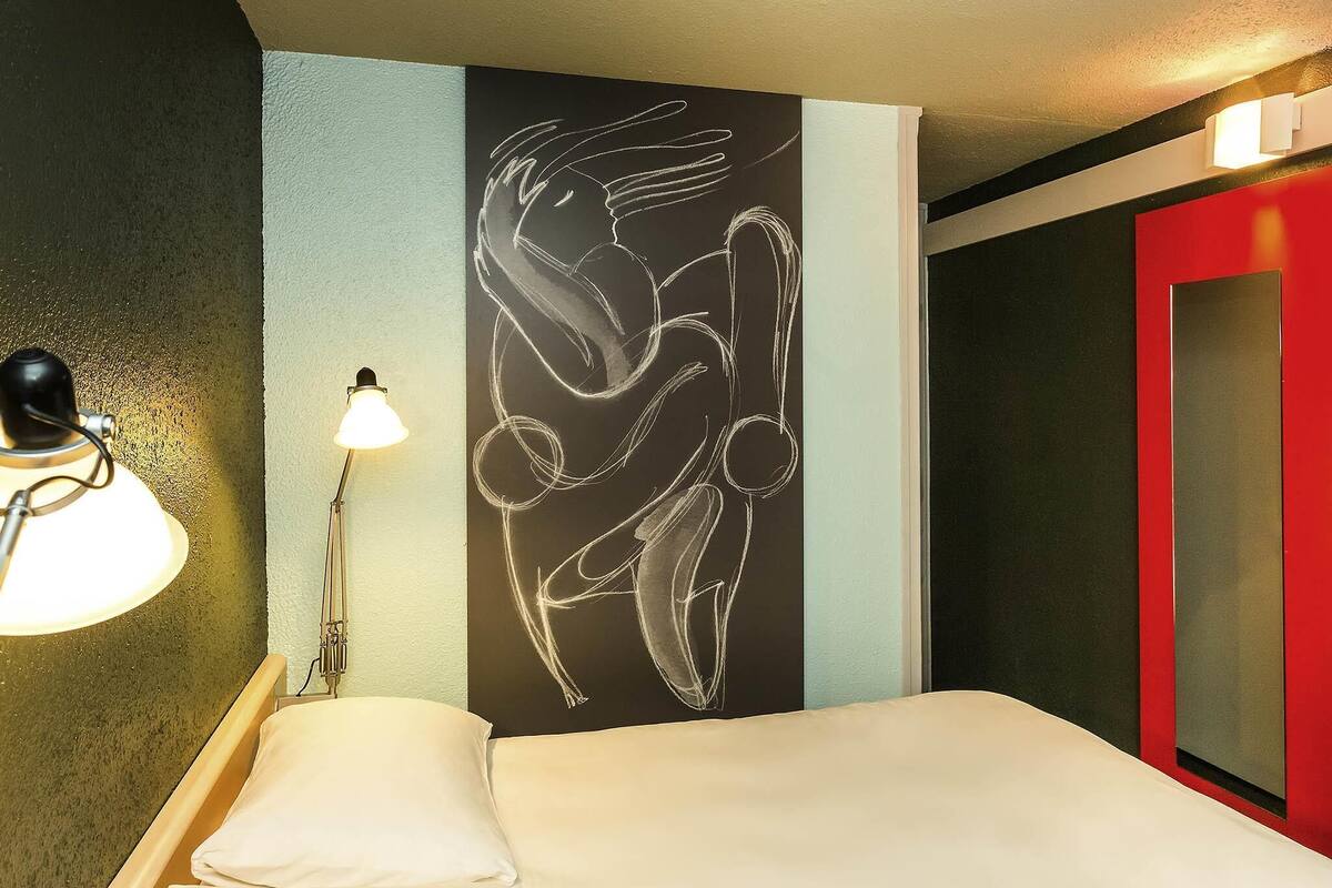 Une chambre d’hôtel avec un lit, une lampe fixée au mur et une porte rouge.