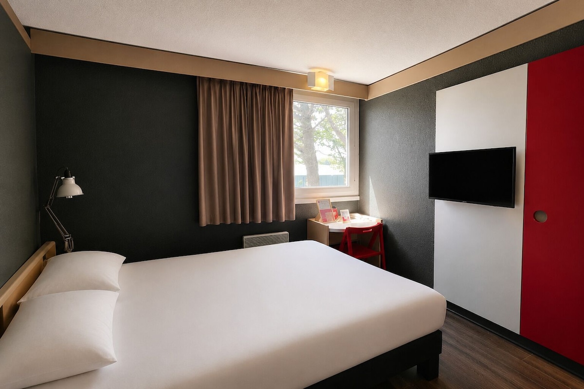 A hotel room with a bed, a desk, a chair, a TV, and a window with curtains.