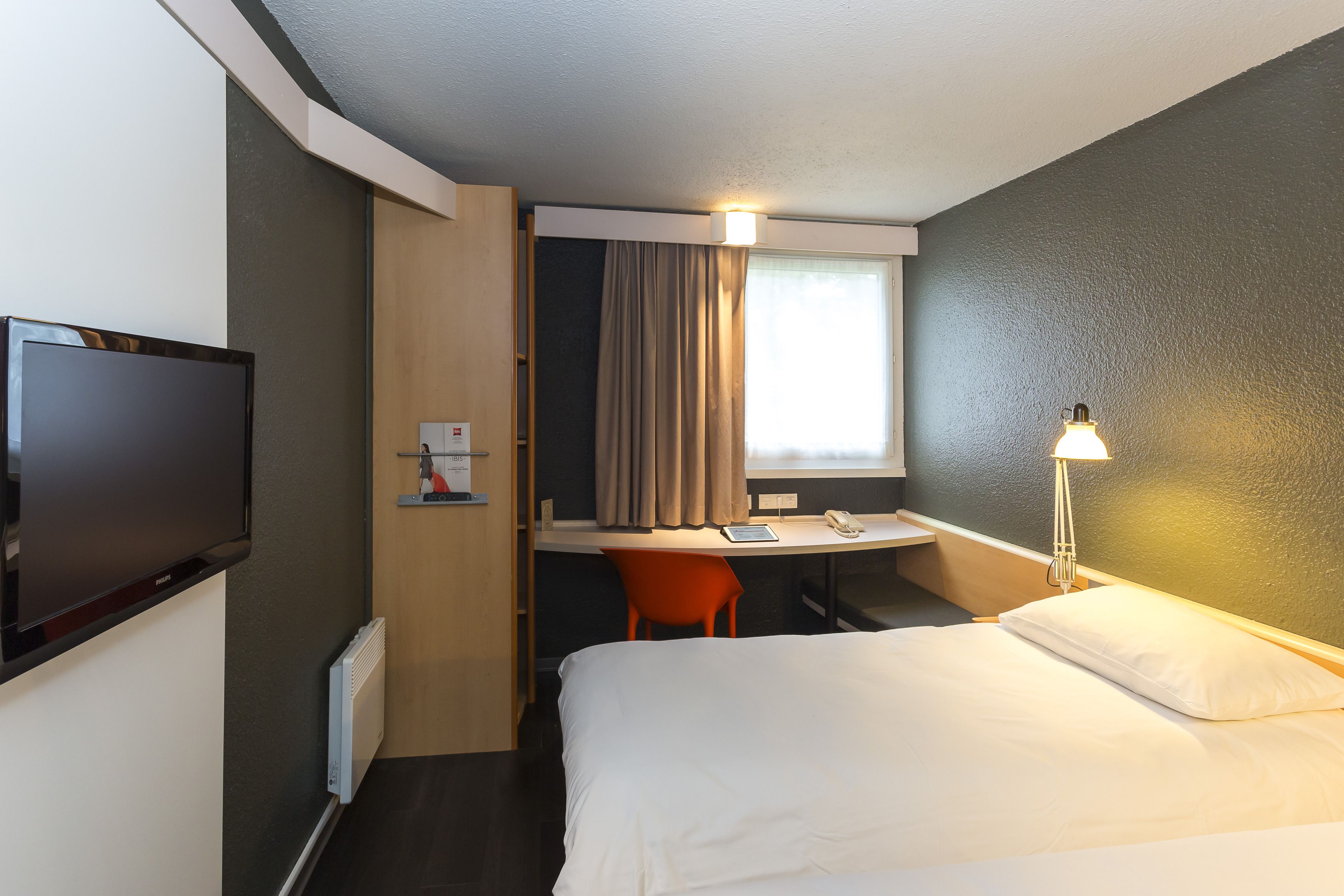 A hotel room with a bed, a desk with a chair, a television, and a window with curtains.