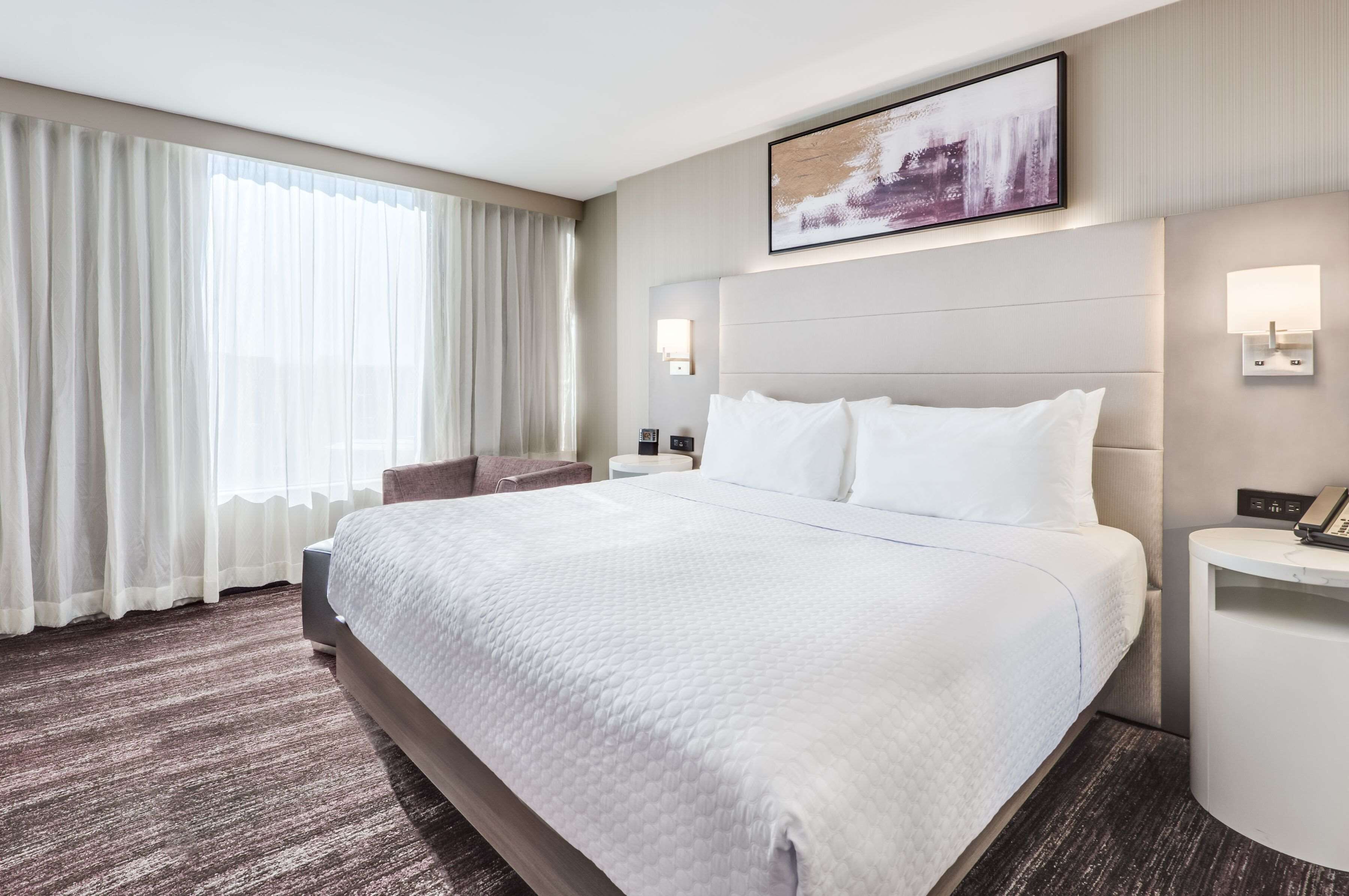 A hotel room with a large bed, a bedside table, a nightstand, a window with curtains, and a wall-mounted artwork.