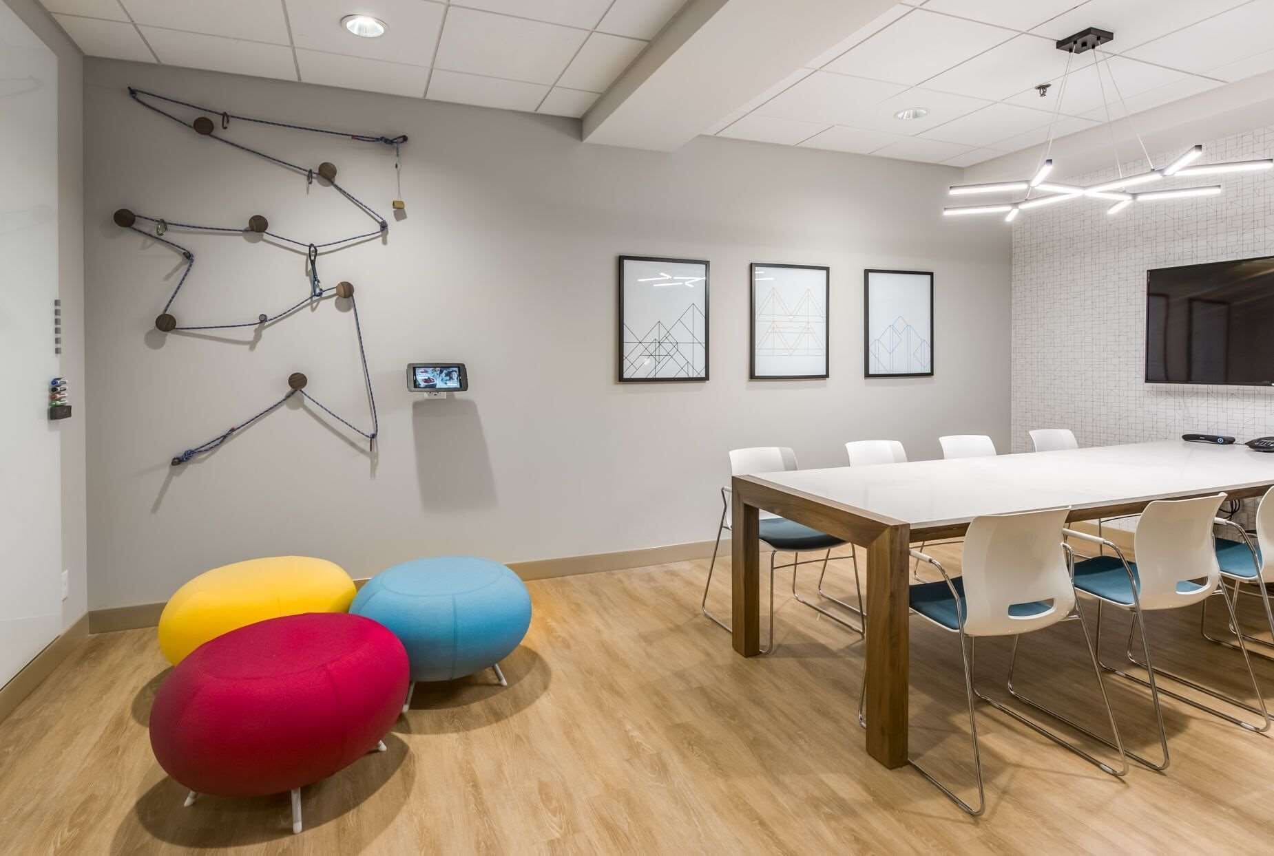 Una sala de reuniones moderna con una mesa blanca, sillas azules y rojas, y arte geométrico en la pared.