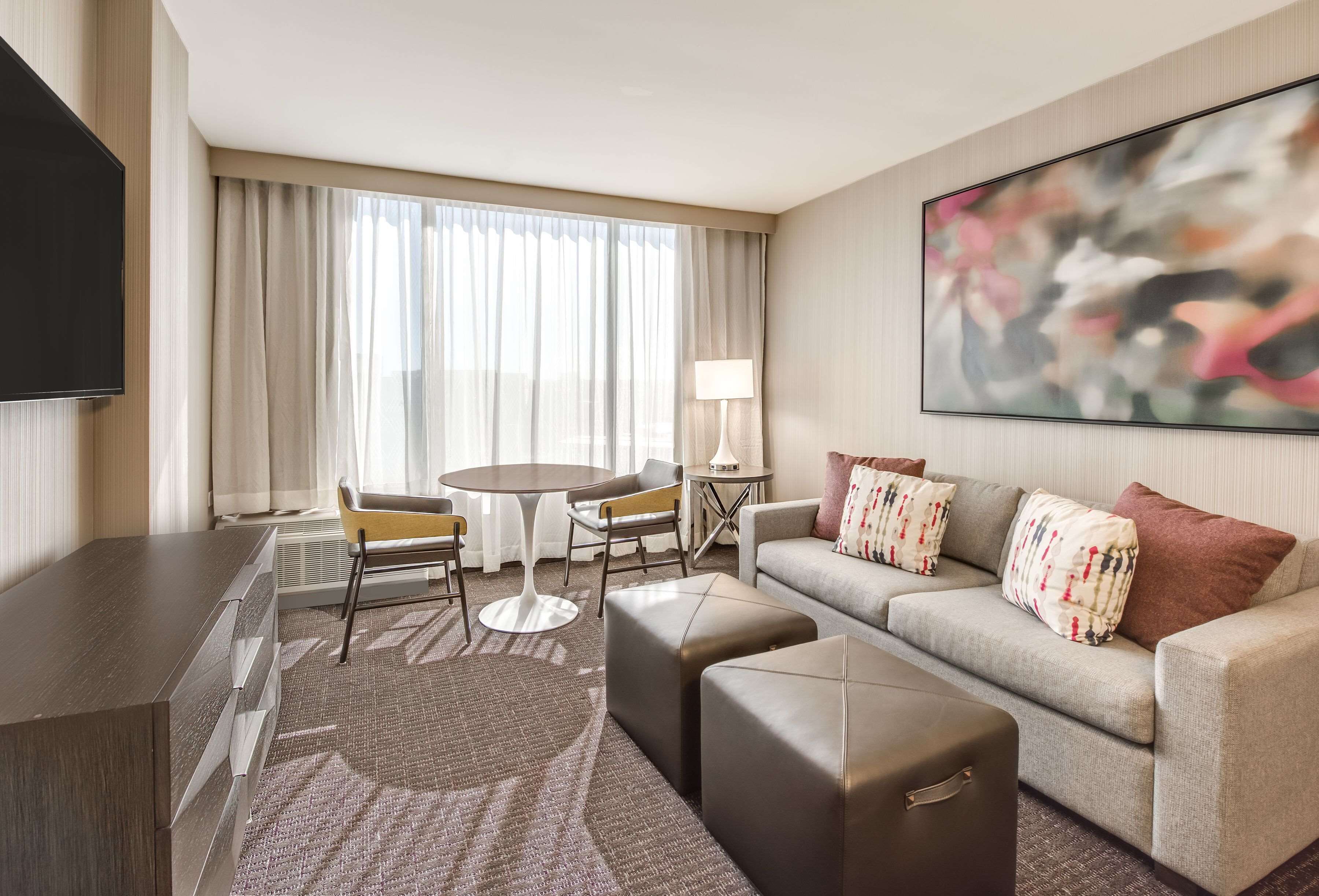 A modern hotel room with a flat-screen TV, a sofa with patterned cushions, a coffee table, and a large abstract painting on the wall.