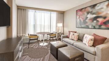A modern hotel room with a flat-screen TV, a sofa with patterned cushions, a coffee table, and a large abstract painting on the wall.