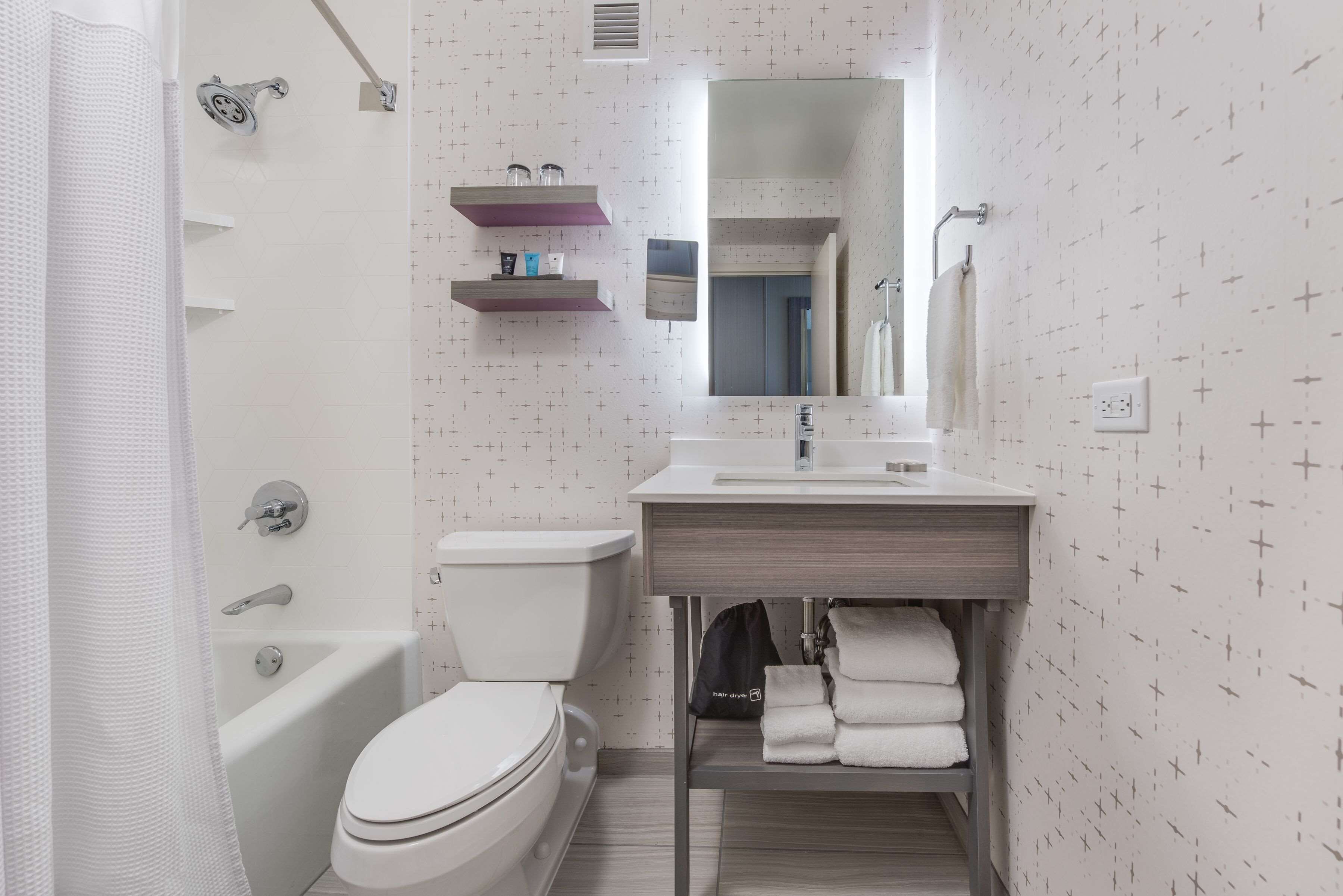 A bathroom with a toilet, bathtub, and a sink with a mirror above it. There are shelves above the sink and a towel rack to the right.