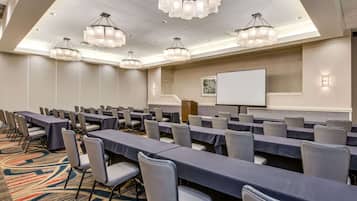A conference room with rows of tables and chairs, a projector screen, and chandeliers.