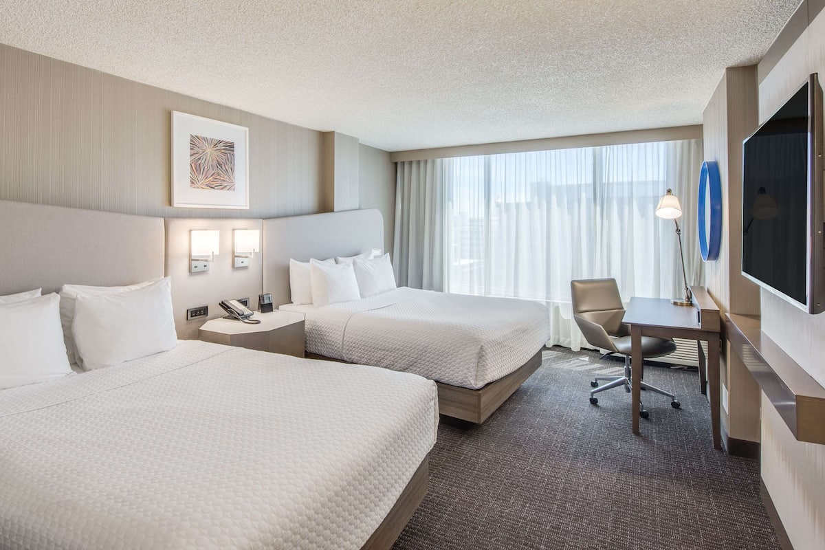 A hotel room with two beds, a desk, a chair, a TV, and a window with curtains.