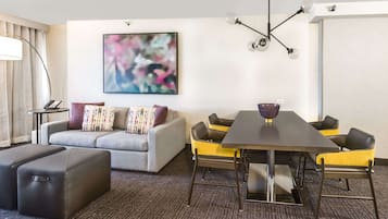 A modern hotel room with a grey sofa, a black ottoman, a dining table with yellow chairs, and a large abstract painting on the wall.