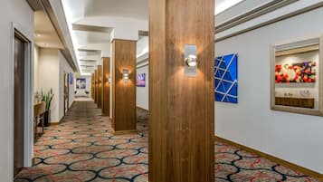 A hotel hallway with patterned carpet, wooden pillars, wall-mounted light fixtures, and artwork.