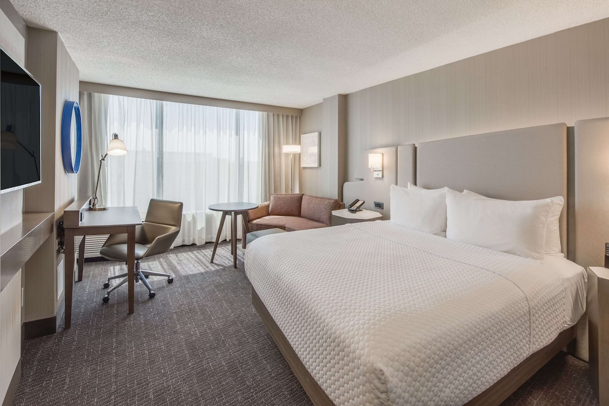 A hotel room with a large bed, a desk with a chair, a small table, a sofa, and a TV mounted on the wall.