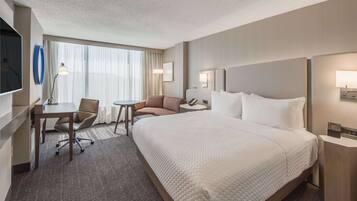 A hotel room with a large bed, a desk with a chair, a small table, a sofa, and a TV mounted on the wall.