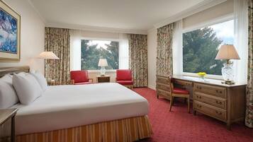 A hotel room with a large bed, a desk, a chair, and a window with a view of trees.