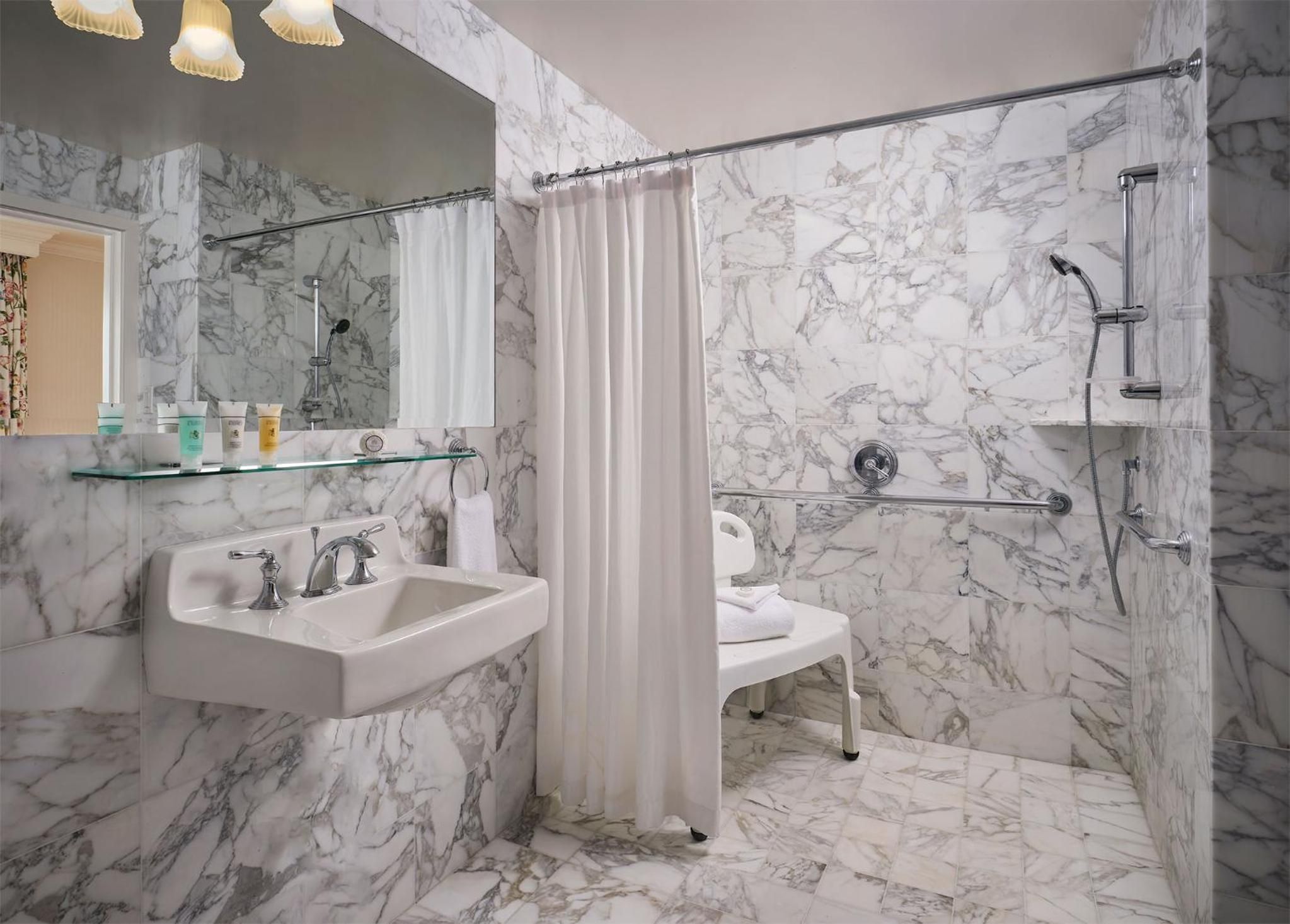 A bathroom with marble walls, a glass shelf with toiletries, a white sink, and a shower area.