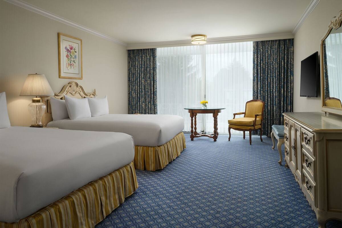 A hotel room with two beds, a small table with a vase, a chair, a TV, and a dresser.