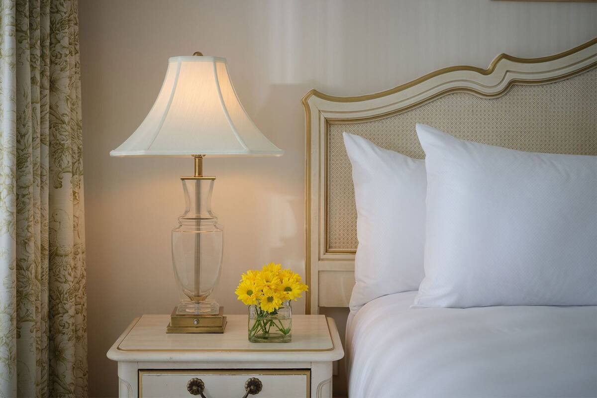 A bed with white bedding, a nightstand with a lamp, and a vase with yellow flowers.