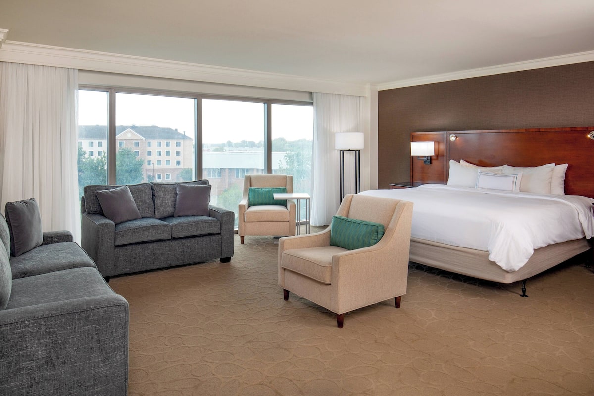 A hotel room with a large bed, two armchairs, a sofa, and a view of a building through large windows.
