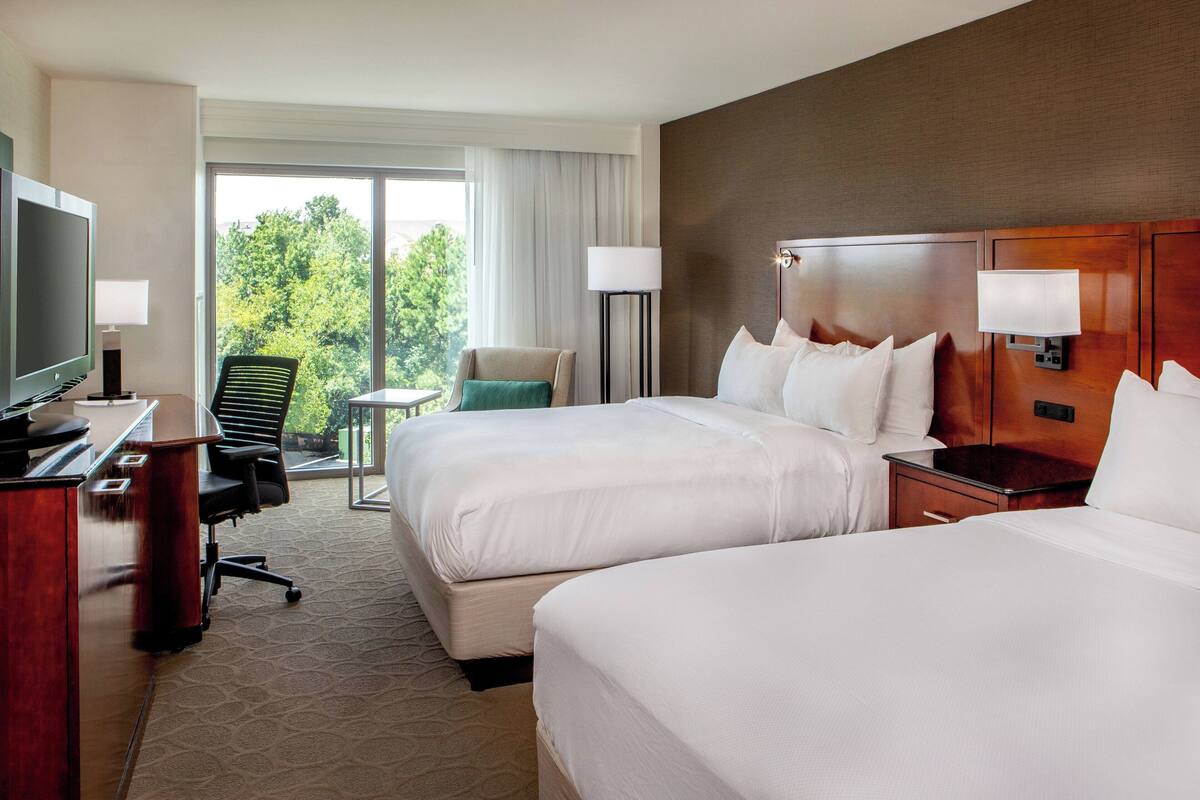 A hotel room with two beds, a desk with a television, a chair, a small table, and a view of trees through the window.