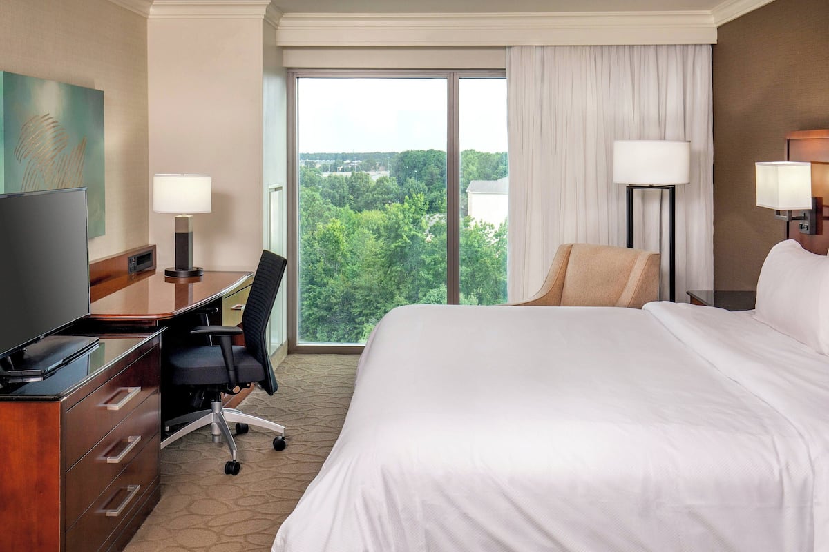 A hotel room with a large bed, a desk with a computer, a chair, and two lamps. There is a large window with a view of trees and buildings.