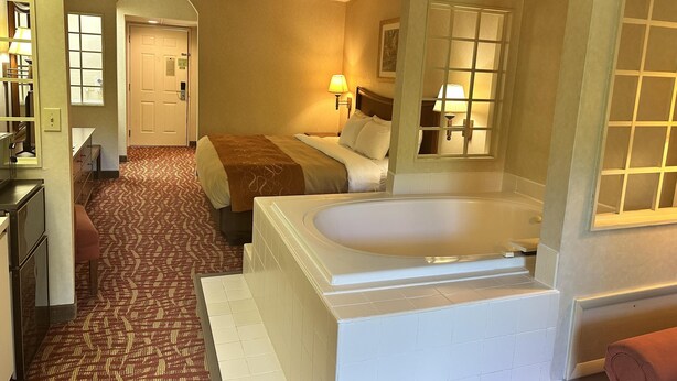A hotel room with a large bathtub, a bed with a brown blanket, a patterned carpet, and a hallway leading to another room. - SureStay by Best Western Cherokee on the River (Cherokee)