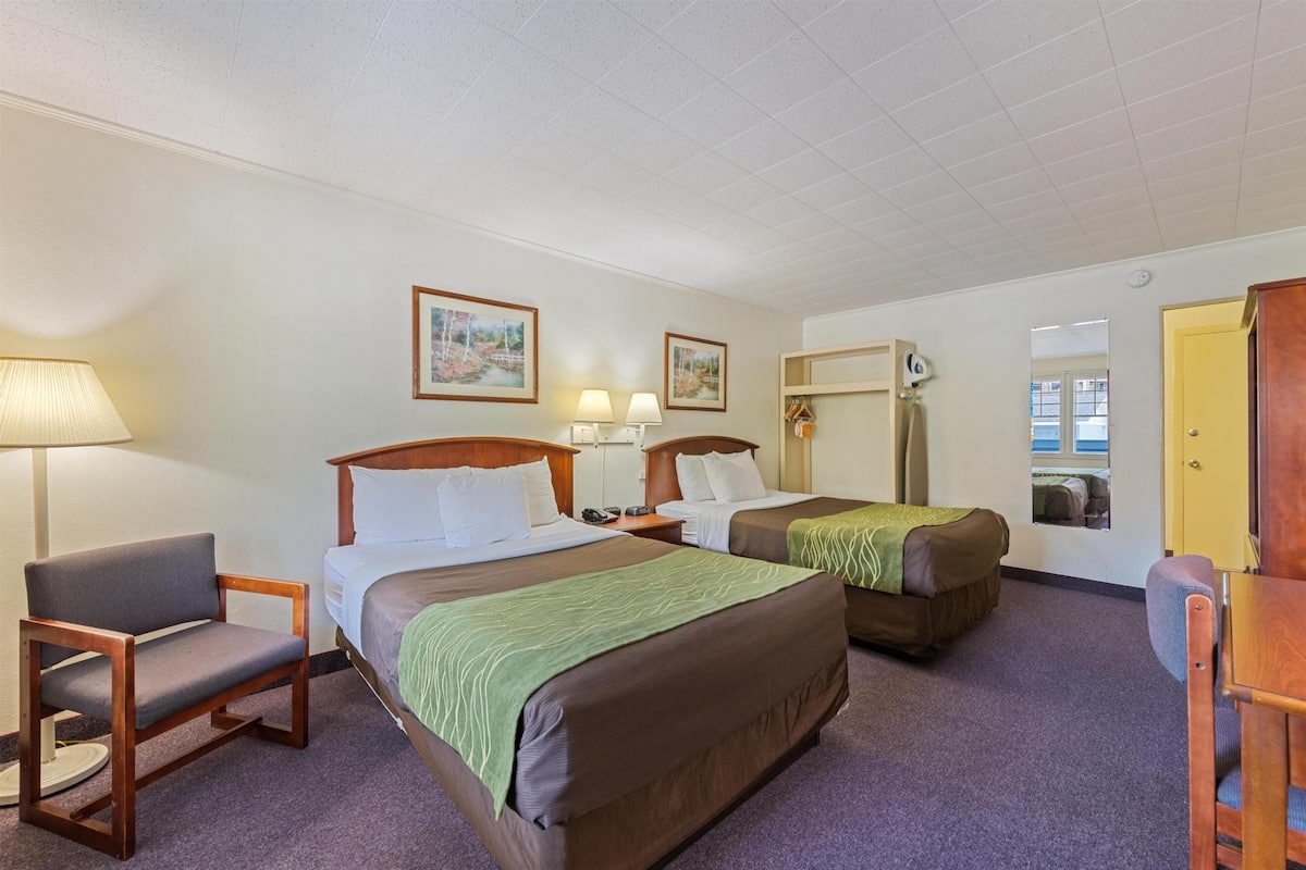 Quarto de hotel com duas camas, uma cadeira, uma escrivaninha, um abajur e um espelho.