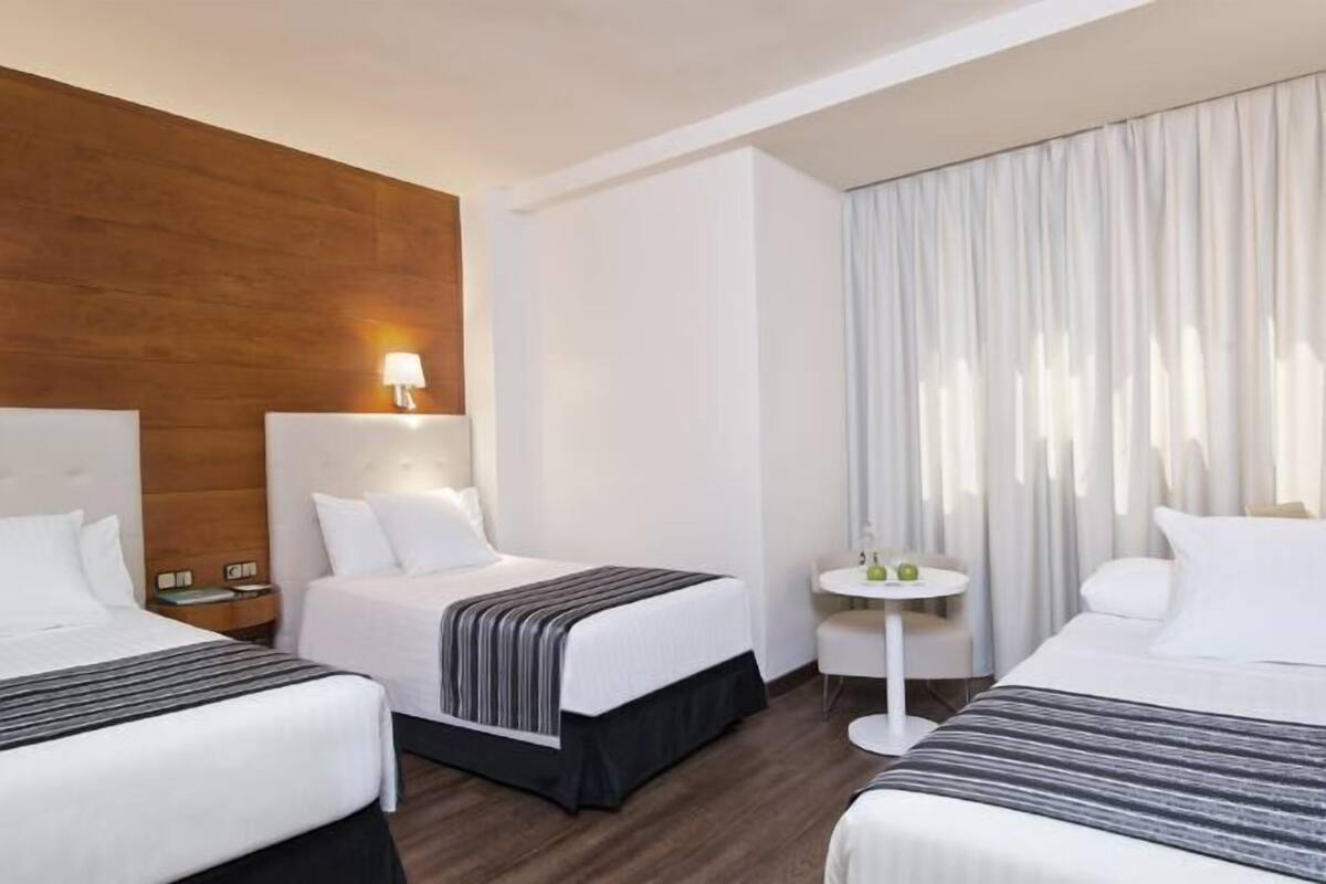 A hotel room with two beds, a small table, a lamp, and a view of the outdoors.