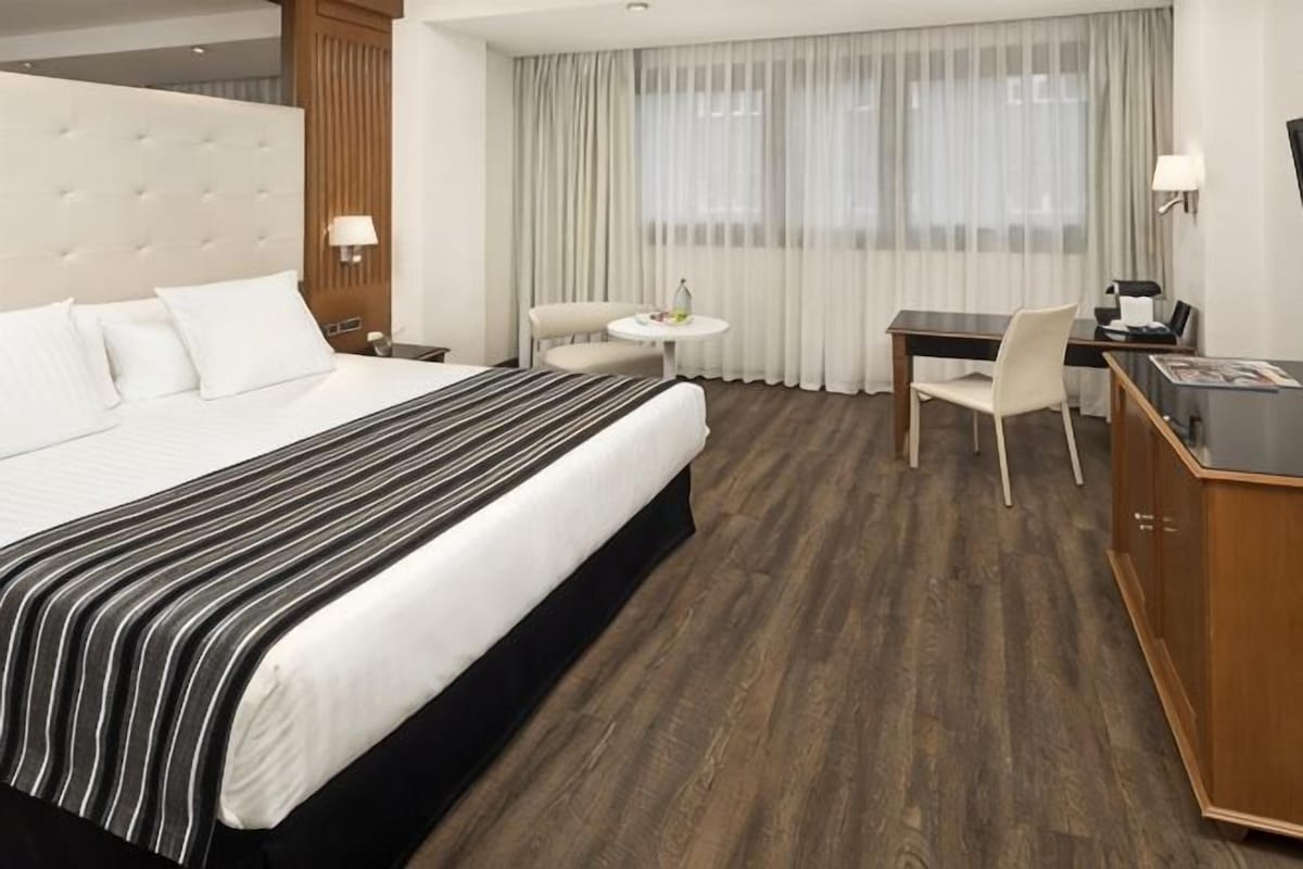 A hotel room with a large bed, a desk, a chair, and a small table.