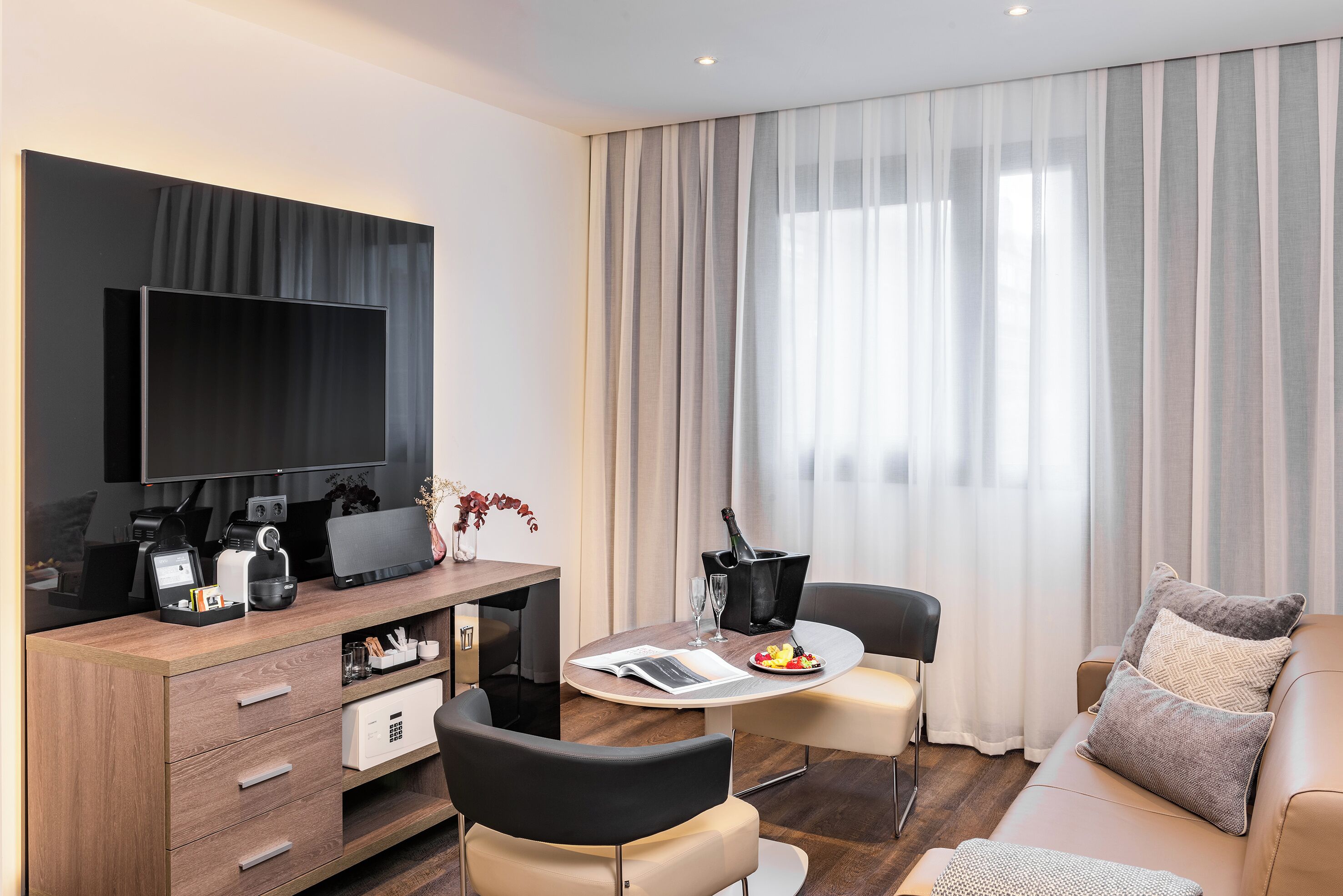 Una habitación de hotel moderna con televisor de pantalla plana, escritorio de madera, zona de comedor con mesa y sillas, sofá y ventana grande con cortinas.