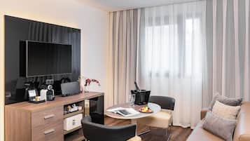 Una habitación de hotel moderna con televisor de pantalla plana, escritorio de madera, zona de comedor con mesa y sillas, sofá y ventana grande con cortinas.