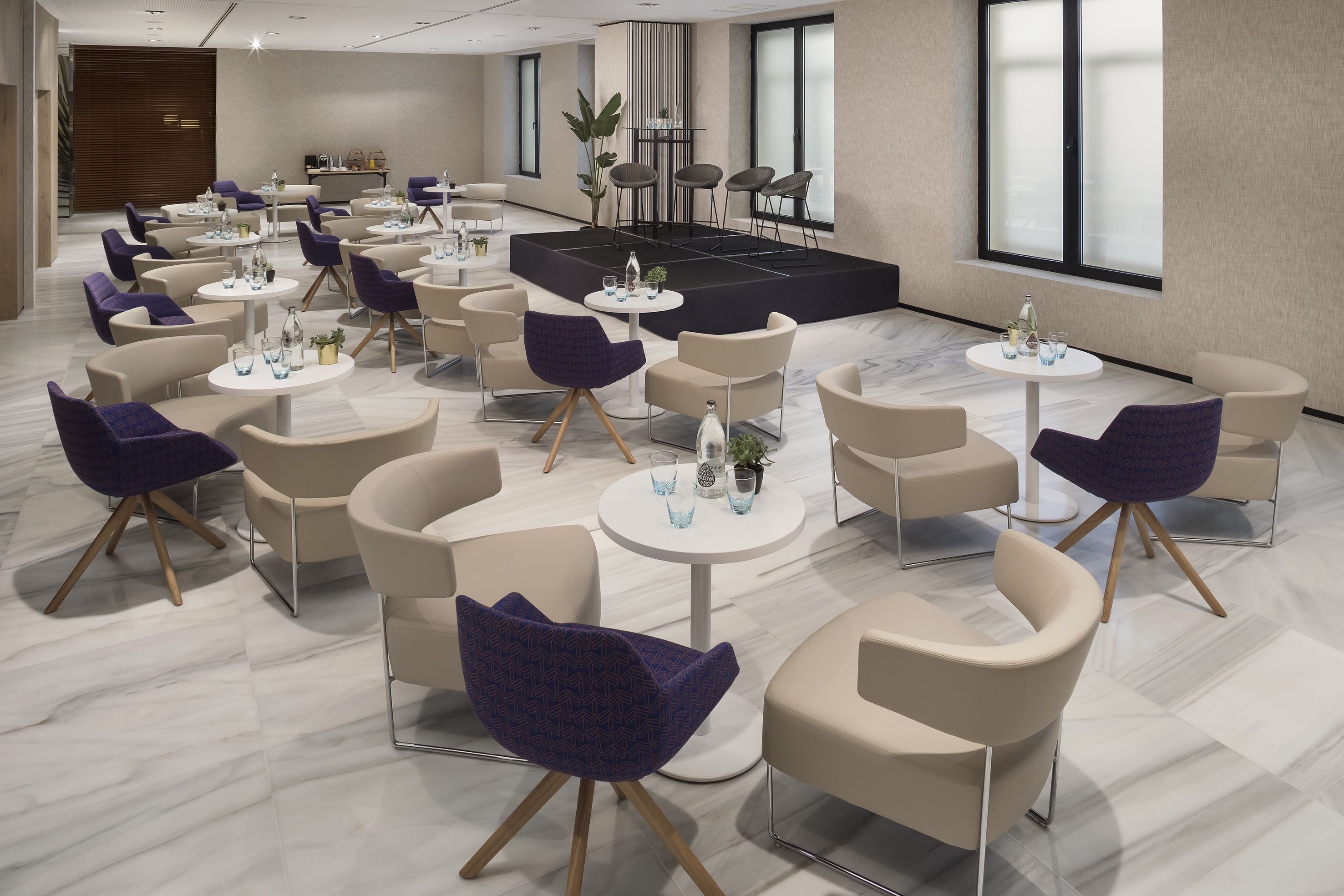 A modern hotel lobby with round tables, comfortable seating, a stage, and large windows.