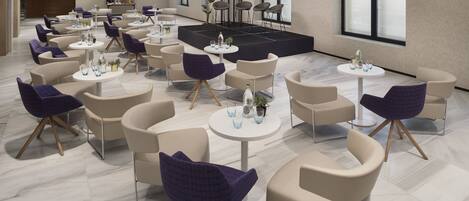 A modern hotel lobby with round tables, comfortable seating, a stage, and large windows.