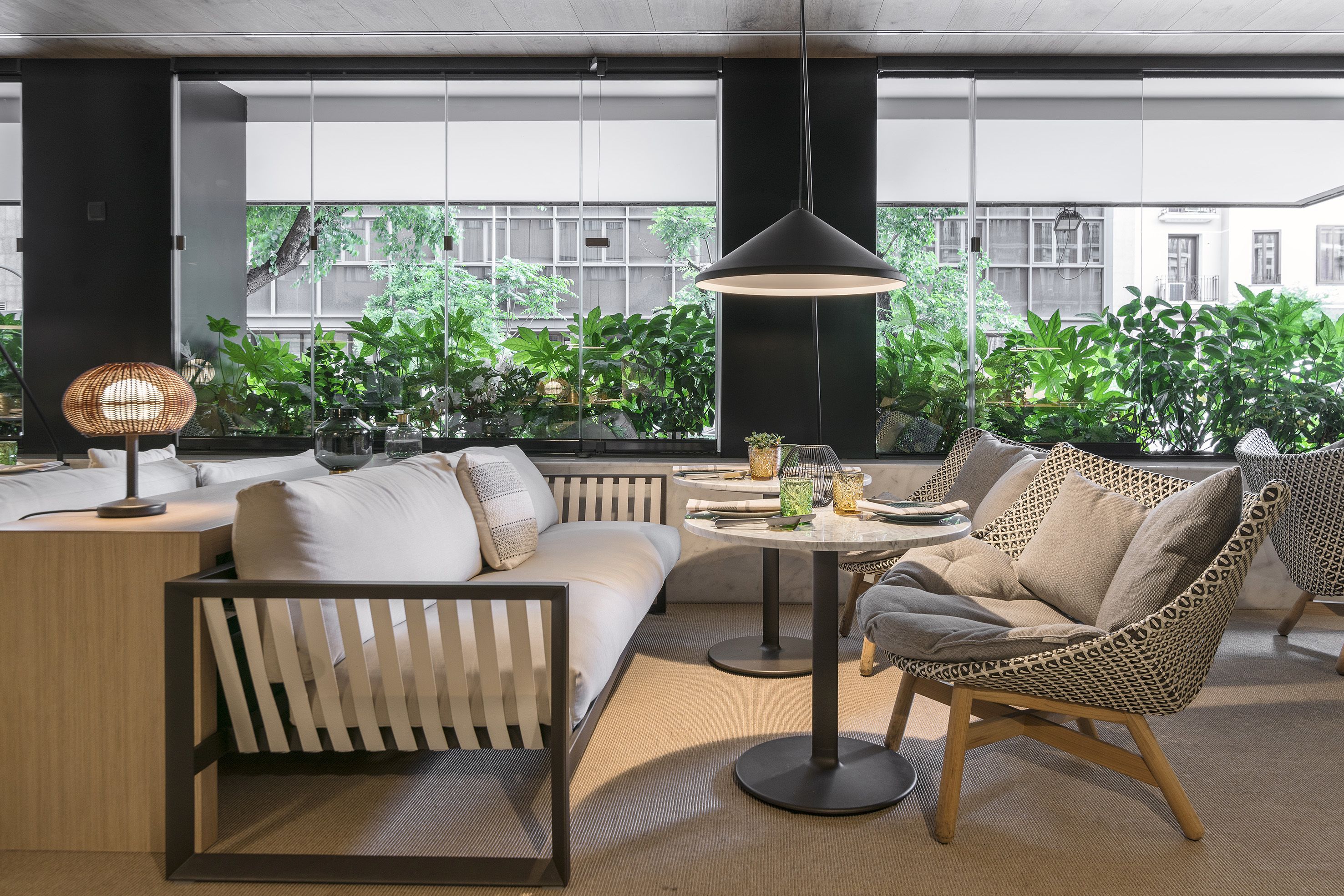 Un área de estar moderna con un sofá, un sillón estampado y una mesa con lámpara. Amplias ventanas ofrecen vistas a la vegetación exterior.
