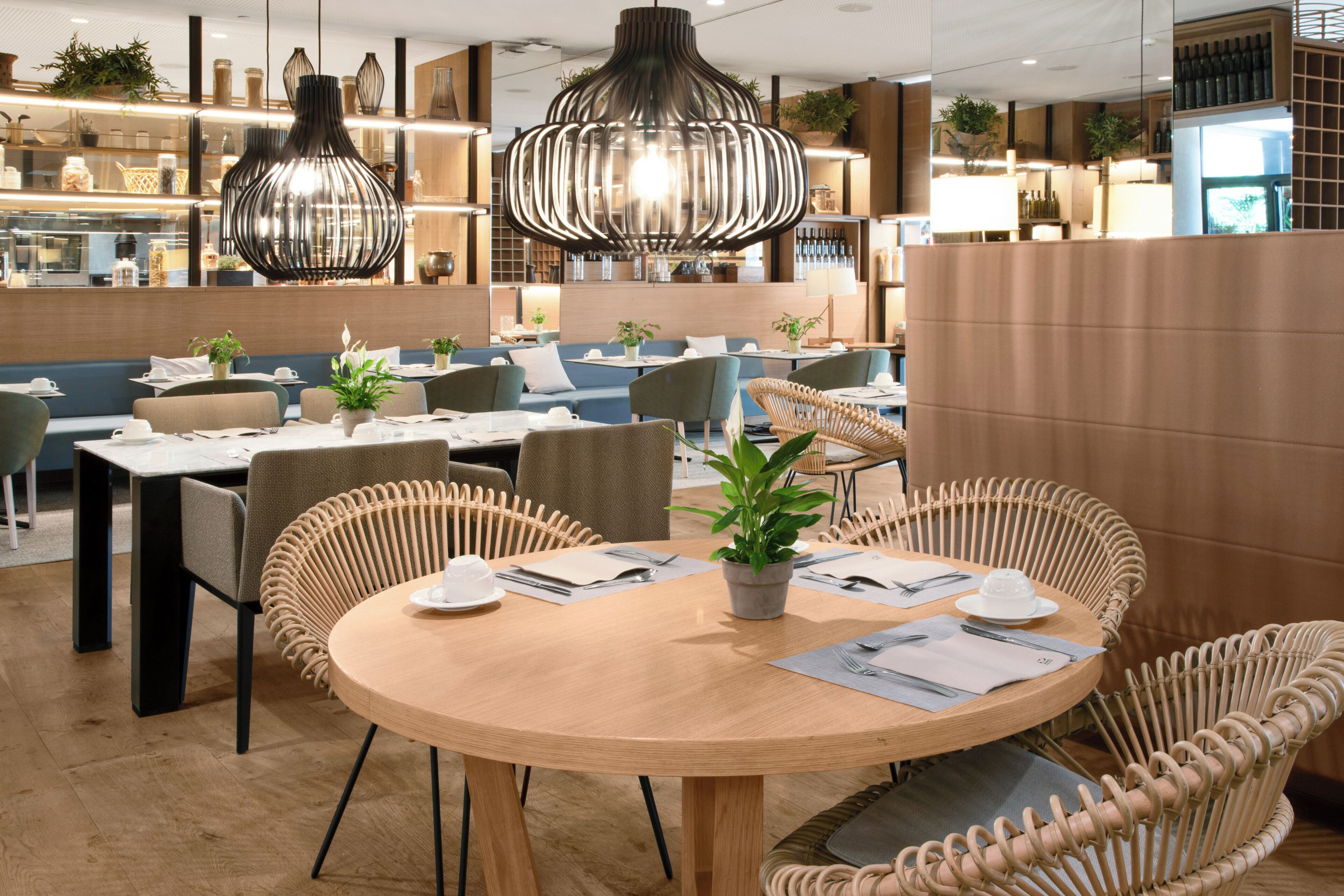 A modern restaurant with round tables, wicker chairs, and hanging pendant lights.